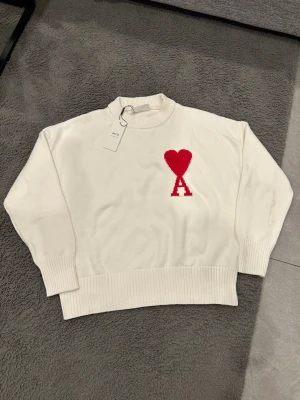 Vit stickad tröja från Ami - Säljer en vit stickad tröja från Ami med röd hjärtlogga och bokstaven A på bröstet. Tröjan har ribbade muddar, rund hals och är långärmad. Perfekt för dig som gillar stilrena plagg med en twist.