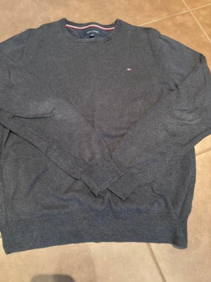 Mörkgrå stickad tröja Tommy Hilfiger - Säljer en mörkgrå stickad tröja från Tommy Hilfiger i storlek L. Tröjan har rund halsringning, diskret logga på bröstet och ribbade muddar vid ärmslut och nederkant. Tillverkad i 100% bomull, perfekt för höst och vinter.