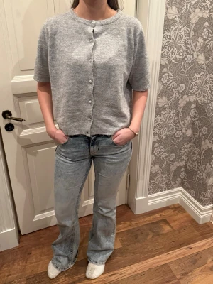 Grå kofta med pärlknappar - Kortärmad grå kofta i mjukt stickat material med pärlknappar framtill. Klassisk rund halsringning och ribbade kanter. Perfekt att slänga över en topp för en chill look. Sista knappen är tyvärr borta.