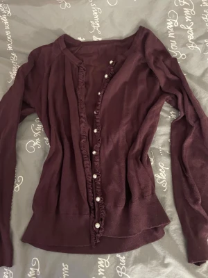 Mörklila kofta med pärlknappar - Säljer en mörklila kofta med långa ärmar och fina volangdetaljer längs knappraden. Koftan har pärlknappar framtill som ger en elegant touch. Materialet känns mjukt och lätt, perfekt för lager på lager under höst och vinter.