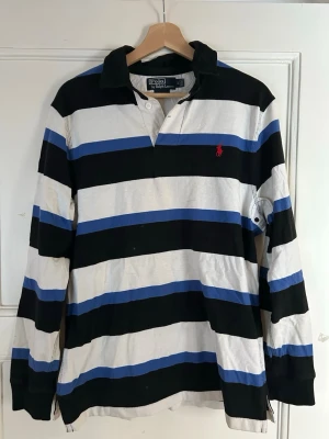 Randig långärmad piké Polo Ralph Lauren - Säljer en klassisk långärmad skjorta från Polo Ralph Lauren i storlek M. Skjortan har breda ränder i svart, vit och blå, med röd broderad logga på bröstet. Tillverkad i mjuk bomull och har en krage med knappslå framtill. Perfekt för dig som gillar sportig och stilren look.