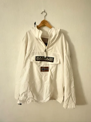 Vit vindjacka från Napapijri - Säljer en vit vindjacka från Napapijri med huva och halv dragkedja. Jackan har en stor ficka framtill med logotyp och norsk flagga, samt en snygg patch på ärmen. Materialet är polyester och jackan är långärmad med loose passform.
