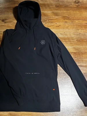 Svart vindjacka från Dope, strl M - Svart vindjacka från Dope med huva och snörning med orange detaljer. Jackan har en stor magficka med tryck och stilren logga på bröstet. Tillverkad i polyester, perfekt för blåsiga dagar. Långärmad och loose fit för skön känsla.