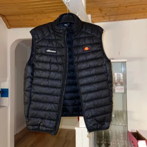 Svart dunväst från Ellesse - Säljer en svart dunväst från Ellesse. Västen har dragkedja framtill och två sidofickor. Köpt för ungefär 3 årsen, använd väl. Den ser fortfarande ny ut bara att den har fått 3 bränn märken på baksidan av västen som man ser på sista bilden. Passformen och längd finns på sista bilden. Ordinarie pris 1 095kr mitt pris 200kr, Priset går att diskutera 🌸