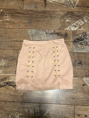 Beige minikjol med snörning  - Säljer en beige kortkjol från DM Retro i mjuk mockaliknande polyester. Kjolen har snygga snörningsdetaljer framtill och två små slitsar längst ner. Dragkedja bak för enkel på- och avtagning. Perfekt för dig som gillar trendiga detaljer och neutral färg.
