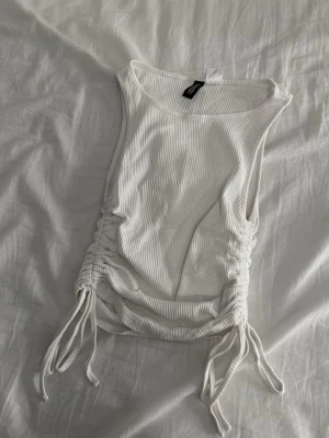 Vit ribbad linnetopp från H&M Divided - Säljer en vit ribbad linnetopp från H&M Divided i storlek XS. Toppen är ärmlös och har snygga snörningsdetaljer på båda sidorna som ger en cool och justerbar passform. Perfekt för varma dagar och enkel att matcha med jeans eller kjol.