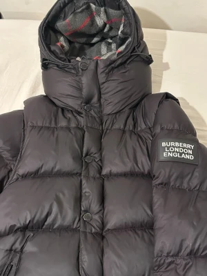 Svart pufferjacka från Burberry - Svart pufferjacka från Burberry London England med avtagbara ärmar så den kan användas som väst. Jackan har huva med rutigt foder i grått och rött, knappar framtill och dragkedja i fickan. Perfekt för kalla dagar och har en riktigt schysst streetstil. Ett fel me vänster ficka på dragkedjan.