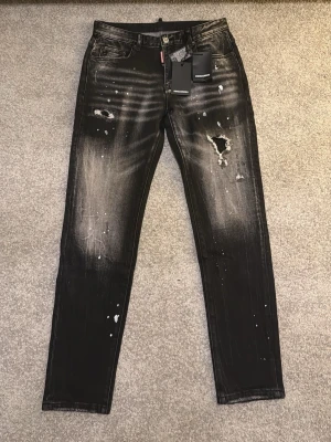Dsquared2 jeans - Säljer ett par svarta jeans från Dsquared2 med coola slitningar, färgstänk och distressed detaljer. Jeansen har en smal passform och klassisk femficksdesign. Materialet är jeans med en tvättad look och läderpatch bak i midjan.