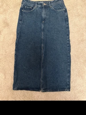 Mörkblå jeanskjol med fickor - Säljer en klassisk mörkblå lång jeanskjol med raka linjer och fem fickor, inklusive två bakfickor. Kjolen har hällor för bälte och knappstängning fram. Perfekt för dig som gillar enkel och tidlös denimstil.