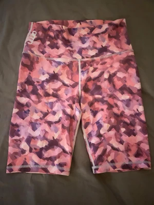 Rosa camo biker sport shorts från Aim'n - Säljer ett par biker shorts från Aim'n i storlek S med rosa och lila camouflage-mönster. De är högmidjade och gjorda i stretchigt material med 73% polyester och 27% spandex, perfekta för träning eller chill. Snygg logga på linningen.