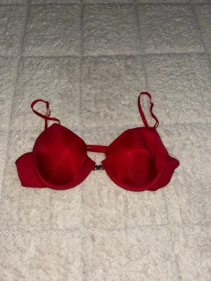 Röd bh Lindex - Snygg röd bh från Lindex med glansigt tyg och hjärtformad metallknäppning framtill. Bh:n har tunna axelband och vadderade kupor för extra stöd. Perfekt för dig som gillar detaljer och vill ha något som sticker ut lite extra.