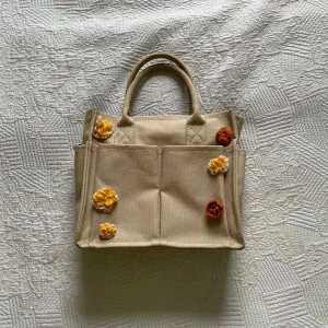 Beige handväska med blommor från Zara - Superfin beige handväska från Zara x Smiley med broderade blommor i gult och oranget. Väskan är i kraftig bomullscanvas, har två handtag, flera fack och dragkedja upptill. Perfekt för dig som vill ha en söt och unik accessoar till vardagen.