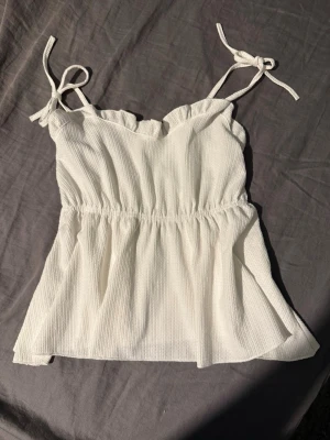 Vit linnetopp med knytband 157 XS - Säljer en söt vit linnetopp från 157 i storlek XS. Toppen har tunna axelband med knyt och en lätt rynkad midja som ger en luftig och avslappnad siluett. Materialet är mjukt och har en diskret struktur. Perfekt för varma dagar och enkel att matcha.