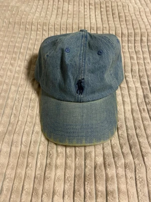 Jeanskeps från Polo Ralph Lauren - Säljer en snygg jeanskeps från Polo Ralph Lauren i klassisk blå denim. Kepsen har broderad logga framtill och justerbart spänne bak. Tillverkad i 100% bomull och har en avslappnad, tidlös look som passar till det mesta.