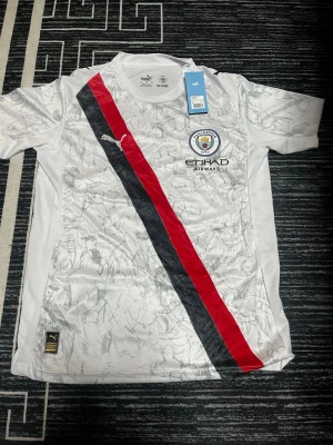 Manchester City fotbollströja Puma M - Säljer en Manchester City fotbollströja från Puma i storlek M. Tröjan är vit med subtilt mönster, har en diagonal röd och svart rand på framsidan, klubbmärke och sponsortryck. På ryggen står det 'Marmoush' och nummer 7. Materialet är polyester.