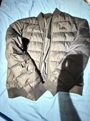 Svart pufferjacka från J.Lindeberg - Säljer en svart pufferjacka från J.Lindeberg med diskret logga på bröstet. Jackan har ribbade muddar, dragkedja framtill och quiltad design. Perfekt för kyliga dagar och ger en clean look till din outfit. Jackan är vändbar till en mer Höst/Vårig look med men passar även till vintern. Aldrig använd. Den är svart på bild men är mörkblå egentligen. 