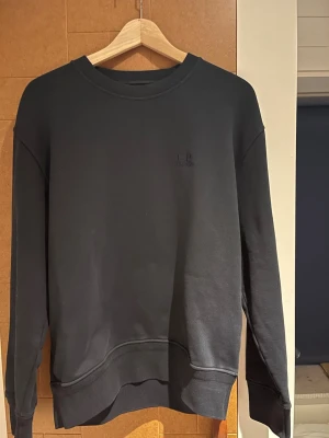 Svart sweatshirt från C.P. Company - En stilren mörkblå sweatshirt från C.P. Company med diskret broderad logga på bröstet. Klassisk rund halsringning och långa ärmar. Perfekt för dig som gillar enkel och clean streetwear. 