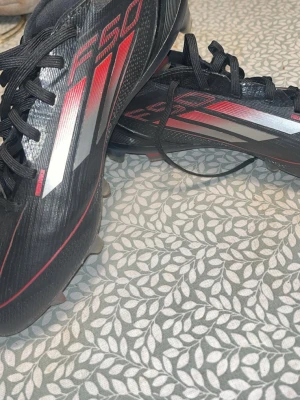 Adidas F50 svarta fotbollsskor - Säljer ett par Adidas F50 fotbollsskor i svart med röda och silvriga detaljer. Skorna har snörning, platt sula med fasta dobbar och är tillverkade i syntetmaterial. Perfekta för dig som vill ha lätta och snabba skor på planen.