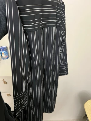 Svart-vit randig kappa - Säljer en svart kappa med vita tunna ränder. Klänningen har långa ärmar, bälte i midjan och slits i sidan. Snygg och stilren design som passar perfekt till dig som gillar en klassisk look.