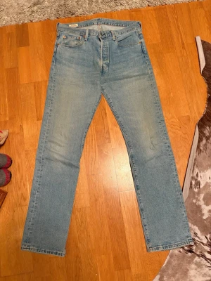 Levi's 501 ljusblå jeans W32 L34 - Säljer ett par klassiska Levi's 501 jeans i ljusblå tvätt med raka ben och knappgylf. Jeansen har fem fickor, bruna detaljer och den ikoniska läderpatchen bak. Perfekt för dig som gillar tidlös denimstil och vill ha ett par jeans med snygg passform.