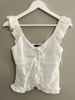 Vit spetsblus med snörning från Bikbok - Supersöt vit blus från Bikbok i storlek S, med broderade blommor och spetsdetaljer på axelbanden. Blusen har snörning framtill, volangkant nedtill och är ärmlös med breda axelband. Perfekt för sommarens alla tillfällen!