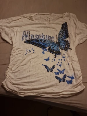 Vit t-shirt med blå fjärilar Lamer&Co - Vit t-shirt från Lamer&Co med stort blått fjärilstryck och flera små fjärilar framtill. T-shirten har korta ärmar och tryckt text på bröstet. Materialet är mjuk bomull med lite stretch för skön passform. Passar nästan vilken storlek som helst. Använd, små synliga deffekter. Priset kan diskuteras🫶