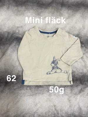 Beige långärmad topp med Disney motiv  - Söt beige långärmad topp med tryckt kaninmotiv i blått nere på framsidan. Toppen har knappar på axeln för enkel påklädning och blå detalj i nederkant. Perfekt för små barn som gillar djurmotiv.🌸 Mini fläck. Storlek 62 Disney kollektion