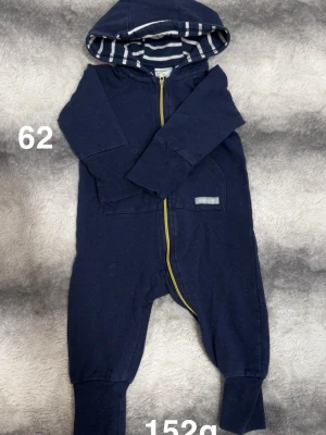 Mörkblå jumpsuit med huva, stl 62 - Mysig mörkblå jumpsuit med huva och gul dragkedja framtill. Insidan av huvan har ett randigt mönster i vitt och blått. perfekt för chill dagar hemma eller när du vill vara bekväm.🌸 Fint använt skick. Storlek 62