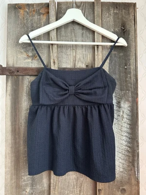 Marinblått linnetopp med rosett - Säljer en marinblå linnetopp med tunna axelband och en stor rosett framtill. Toppen har en lätt rynkad nederdel och är tillverkad i ett mjukt, strukturerat tyg som ger en skön känsla. Perfekt för varma dagar och enkel att matcha med jeans eller kjol.