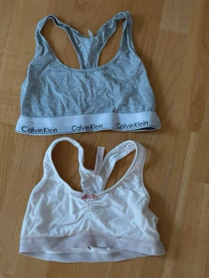Två sportiga bh-toppar från Calvin Klein samt Odd Molly - Säljer två sportiga bh-toppar från Calvin Klein.samt Odd Molly. Ena är ljusgrå med vit resår och logga, den andra är vit med ljusrosa spetsdetalj och liten rosett framtill. Båda har brottarrygg och är perfekta för träning eller chill.
