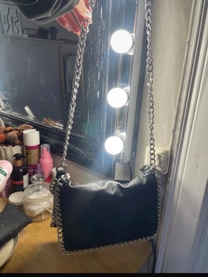 Svart axelväska med kedjedetaljer - Säljer en svart axelväska från TIAMO i skinnimitation med grova silverfärgade kedjor runt kanterna och som axelrem. Väskan har ett huvudfack med dragkedja och en innerficka. Perfekt storlek för det viktigaste och riktigt snygg design.