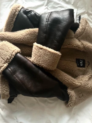 Beige teddyjacka med skinn från Zara - Säljer en beige teddyjacka från Zara med mörkbruna skinnpaneler och fluffigt foder. Jackan har dragkedja framtill och breda muddar i teddy. Perfekt för dig som vill ha en cool och varm look under våren och hösten.