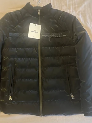 Svart pufferjacka från Moncler - Säljer en svart pufferjacka från Moncler med diskret logga på bröstet. Jackan har två sidofickor med dragkedja och hög krage. Materialet är blankt och känns lyxigt, perfekt för kalla dagar. Dragkedja framtill och stilren design. Storlek S
