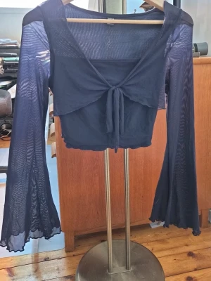 Mörkblå mesh bolerotopp med knyt - Säljer en mörkblå bolerotopp i mesh med långa, utsvängda ärmar och knyt framtill. Toppen är croppad och har en lätt transparent look som ger en cool vibe. Perfekt att styla över ett linne eller klänning för extra edge.