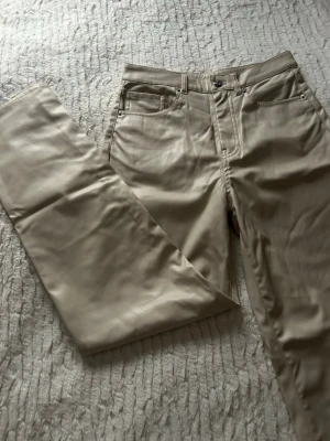 Beiga raka byxor i skinnimitation - Snygga beiga byxor med rak passform och klassisk femficksdesign. Materialet har en skinnliknande finish som ger en cool och modern vibe. Perfekt för dig som vill sticka ut med en stilren look. Byxorna har knapp och dragkedja framtill.