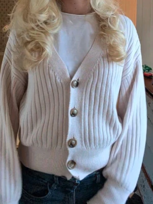Beige ribbad kofta från NA-KD - Säljer en beige ribbad kofta från NA-KD i storlek M. Koftan har v-ringning, fyra knappar framtill och långa ärmar med breda muddar. Materialet är stickat och ger en mjuk och skön känsla. Perfekt att slänga över en t-shirt för en chill look.