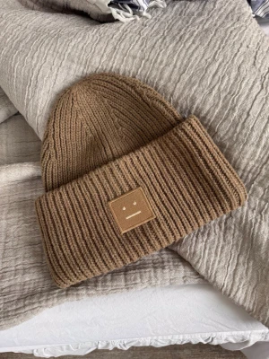 Brun stickad mössa Acne Studios - Säljer en brun stickad mössa från Acne Studios i 100% ull. Mössan har en bred uppvikt kant och Acne Studios klassiska ansiktslogga framtill. Perfekt accessoar för kyliga dagar och riktigt snygg till höst- och vinterlooken. Använd enstaka gång, för tjock för mig