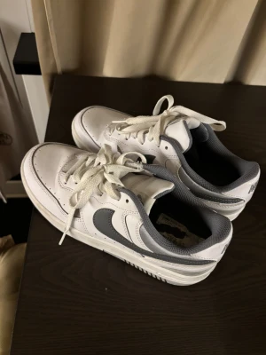 Nike Air Force 1 Low vit/grå - Nike Air Force 1 Low sneakers i vitt och grått med klassisk swoosh på sidan. Skorna har snörning, rund tå och platt sula. Ovandelen är i skinn och mesh, med broderad Nike-logga på hälen. Perfekt för dig som gillar stilrena och tidlösa sneakers. Använt 3 gånger. Storlek 39.