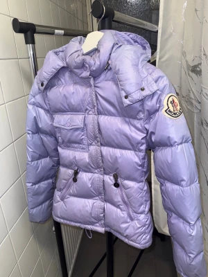Ljuslila Moncler dunjacka gammal design - Säljer en ljuslila dunjacka från Moncler med huva, stora puffiga sektioner, snygga detaljer som dragkedjor och knappar. Jackan har Moncler-logga på ärmen och praktiska fickor framtill. Perfekt för dig som älskar lila o vill ha en superfin vinterjacka. Storlek 1 motsvarande XS/S. Jackan är äkta & köpt från någon här på Plick för några år sen, men har aldrig kommit till användning. Det är en äldre modell med knappar, därför ingen QR kod. Fint skick, bara lite ljusblå färg inuti vid storleksloggan.