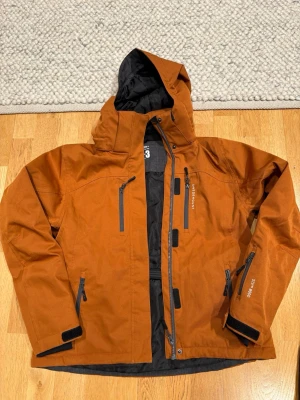 Swedemount orange vindjacka med huva - Säljer en orange vindjacka från Swedemount med flera praktiska fickor med dragkedja och justerbara muddar. Jackan har huva och svart innerfoder, perfekt för utomhusaktiviteter som skidåkning, skridskor mm. Snygg och funktionell design med coola detaljer.