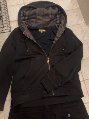 Svart Burberry ZipUp Hoodie - En svart Burberry hoodie med dragkedja som ger din vardagliga outfit en bra touch med det vackra ikoniska burberry mönstret i luvan och insidan på tröjan varför jag säljer den är för att den inte kommer till användning längre och som ni kan se på sista bilden är det några små fläckar vid fickan som jag fått när jag målat med sprayfärg men det vet nog många säkert hur man får bort! Tveka inte på att ställa en fråga eller två❤️ 🔥👍