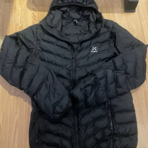 Svart pufferjacka från Haglöfs - Säljer en svart pufferjacka från Haglöfs med huva och dragkedja framtill. 