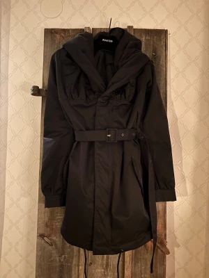 Svart parkajacka från RAERE med bälte och stor krage - Säljer en svart parkajacka från RAERE med stor krage, bälte i midjan (går att ha på två olika ”höjder”) samt snyggt RAERE- märke i paljetter på ena ärmen. Jackan har långa ärmar, dragsko nertill och praktiska fickor. Materialet är polyester och jackan har en clean, oversized look. Den är något nopprig på insidan efter en grå tröja, men absolut inget som stör eller som syns när jackan används!