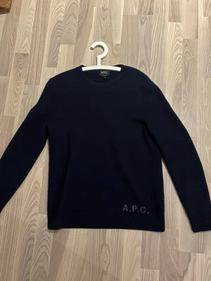 Mörkblå stickad tröja från A.P.C. - Snygg mörkblå stickad tröja från A.P.C. i storlek M. Tröjan har rund hals, ribbade muddar och diskret A.P.C.-logga vid nederkanten. Perfekt för dig som gillar stilrena och klassiska plagg med en modern touch.