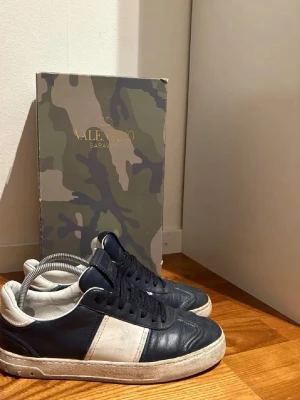 Valentino flycrews - Snygga sneakers från Valentino Garavani i mörkblått och vitt skinn. Klassisk låg modell med rund tå, svarta skosnören och vit platt sula. Kommer med originalkartong i camouflage-mönster. Perfekt för dig som gillar exklusiva märken och stilren design.