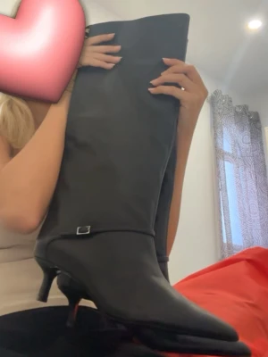 Svarta stövlar med smal och låg klack - Säljer ett par höga svarta stövlar med spetsig tå och smal klack. Stövlarna har en liten metallspänne-detalj vid ankeln och är tillverkade i ett slätt material som ser ut som skinn. Perfekta för dig som vill ha en stilren och edgy look. Endast använda en gång 