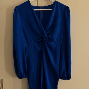 Blå omlottklänning med lång ärm - Snygg blå klänning med omlottdetalj framtill och lång ärm med lätt puff. Klänningen har v-ringad hals och dragkedja i ryggen. Materialet är slätt och har en lätt glans. Perfekt för dig som gillar stilrena och trendiga plagg.