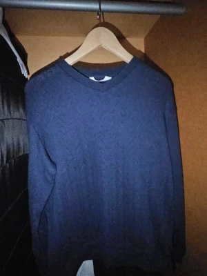 Tröja  - Mörkblå stickad tröja med diskret mönster i tyget. Tröjan har långa ärmar med ribbade muddar och är gjord i ett mjukt material som passar perfekt för kyligare dagar. Enkel och stilren design som funkar till många olika outfits. Helt oanvänd på grund av opassande storlek.
