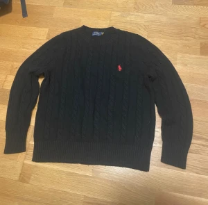Svart kabelstickad tröja Polo Ralph Lauren - Svart kabelstickad tröja från Polo Ralph Lauren med klassisk röd broderad logga på bröstet. Tröjan har rund hals och långa ärmar, perfekt för dig som gillar stilren och tidlös design. Materialet är mjukt och värmande, passar grymt under höst och vinter.