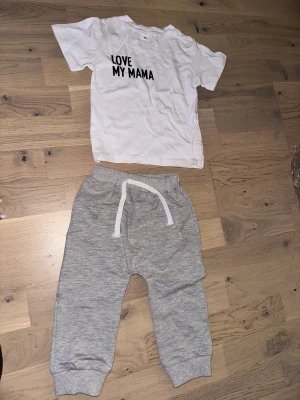 Grå mjukisbyxor med snörning barn Set med T-shirt och Byxor - Supermjuka grå mjukisbyxor för barn med vit snörning i midjan och muddar vid bensluten. Perfekta för chill dagar och lek, tillverkade i bomull för extra komfort.Ett set med en vit t-shirt med texten "LOVE MY MAMA" och ett par grå byxor med dragsko.från 6 mån